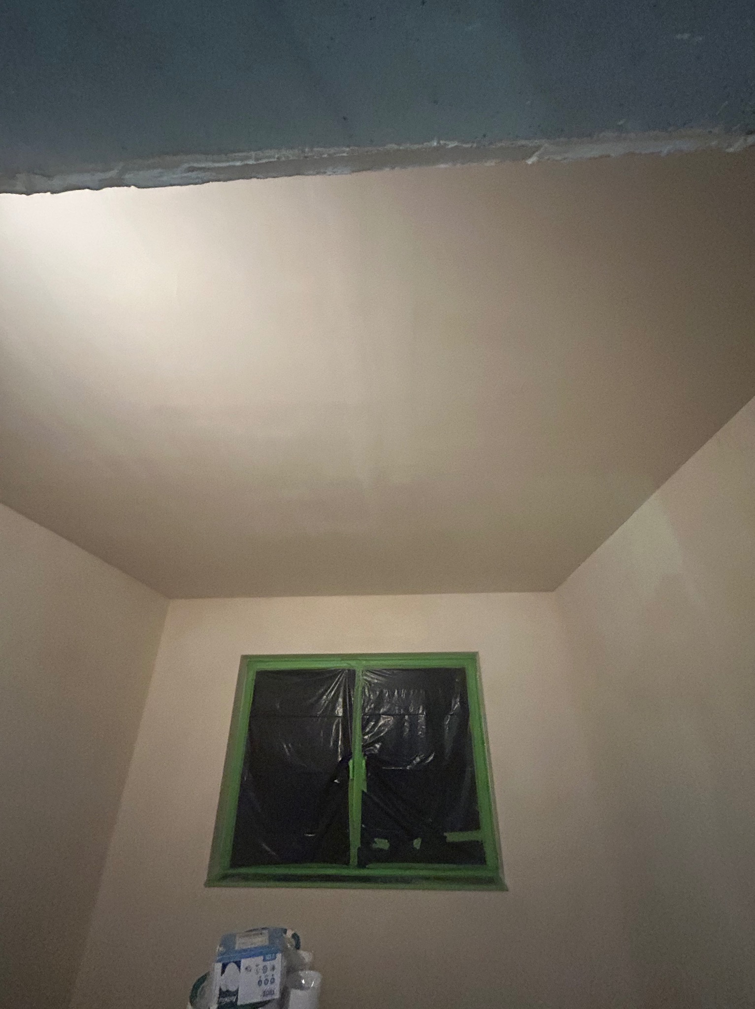 Wygładzony sufit w pokoju z zabezpieczonym oknem folią i taśmą malarską. Widoczne wiaderka z farbą. Świeżo wykończone ściany w jasnym odcieniu.