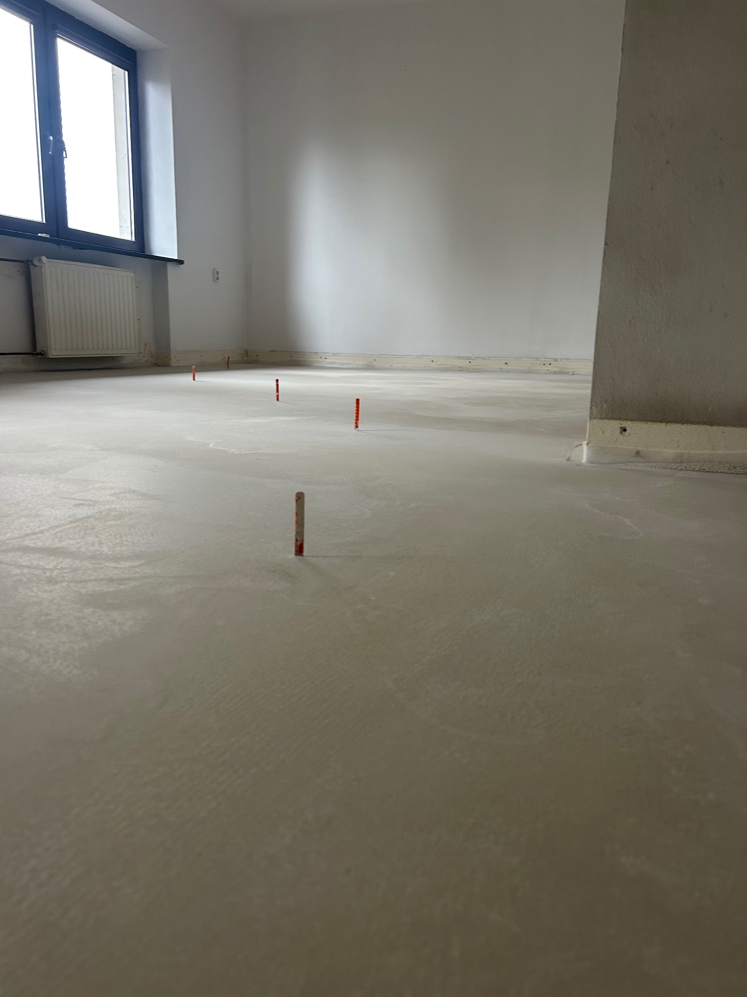 Wnętrze pokoju z wylewką samopoziomującą, widoczne znaczniki wysokości. Ściany białe, okno z grzejnikiem. Podłoga jasnoszara, gładka.