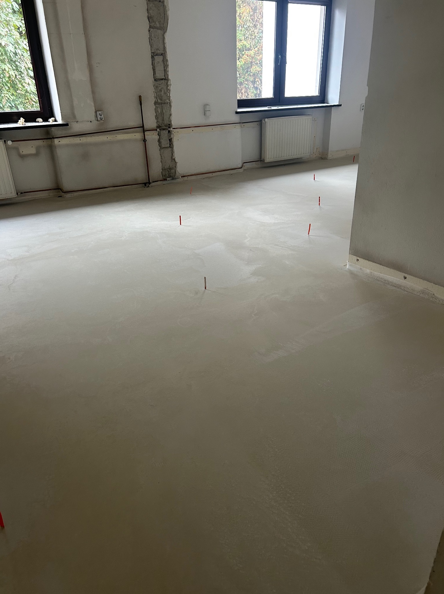 Wylany, świeży podkład podłogowy w jasnym pomieszczeniu z oknami. Widoczne wystające znaczniki poziomu i surowe ściany w trakcie prac wykończeniowych.