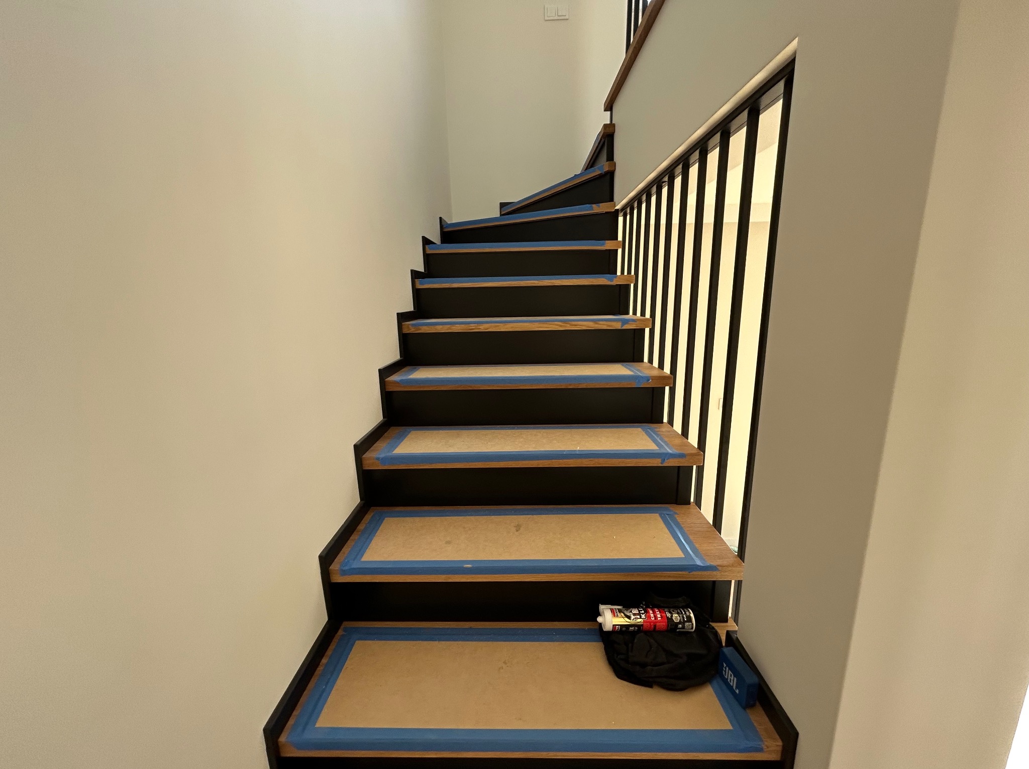 Schody z drewnianymi stopniami zabezpieczone niebieską taśmą malarską przed malowaniem. Widoczne narzędzia i akcesoria malarskie. Ujęcie z dołu, perspektywa w górę.