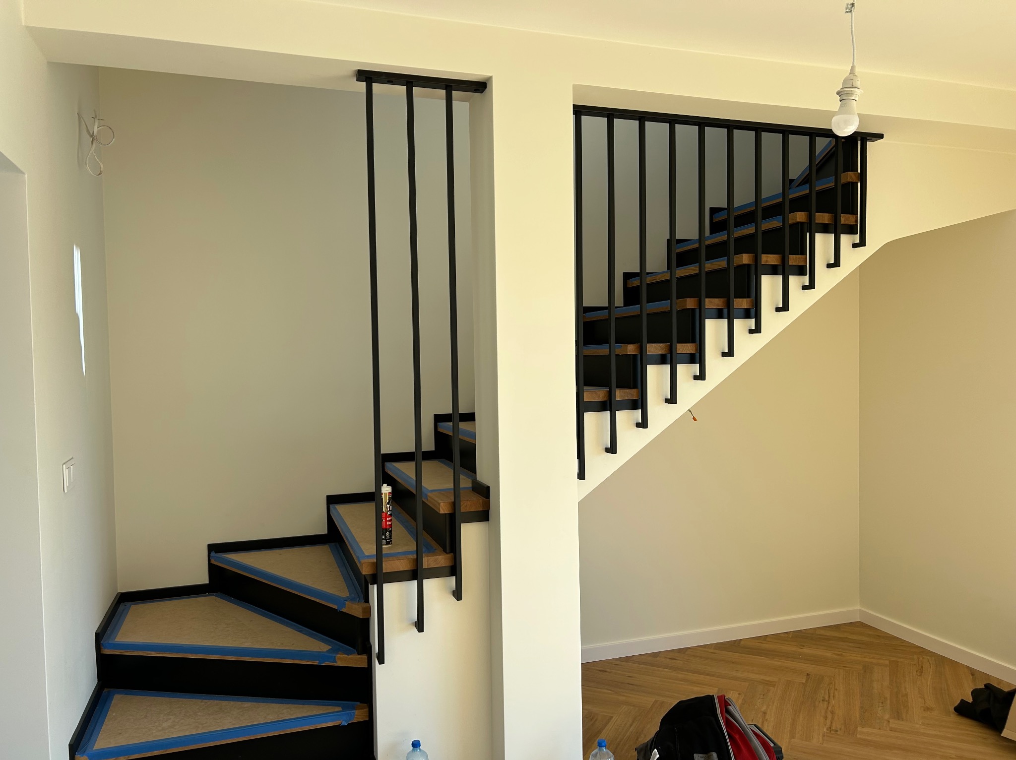 Schody z czarną balustradą w trakcie wykończenia, zabezpieczone taśmą malarską. Widoczna butelka z klejem i podłoga w jodełkę. W tle surowa żarówka.