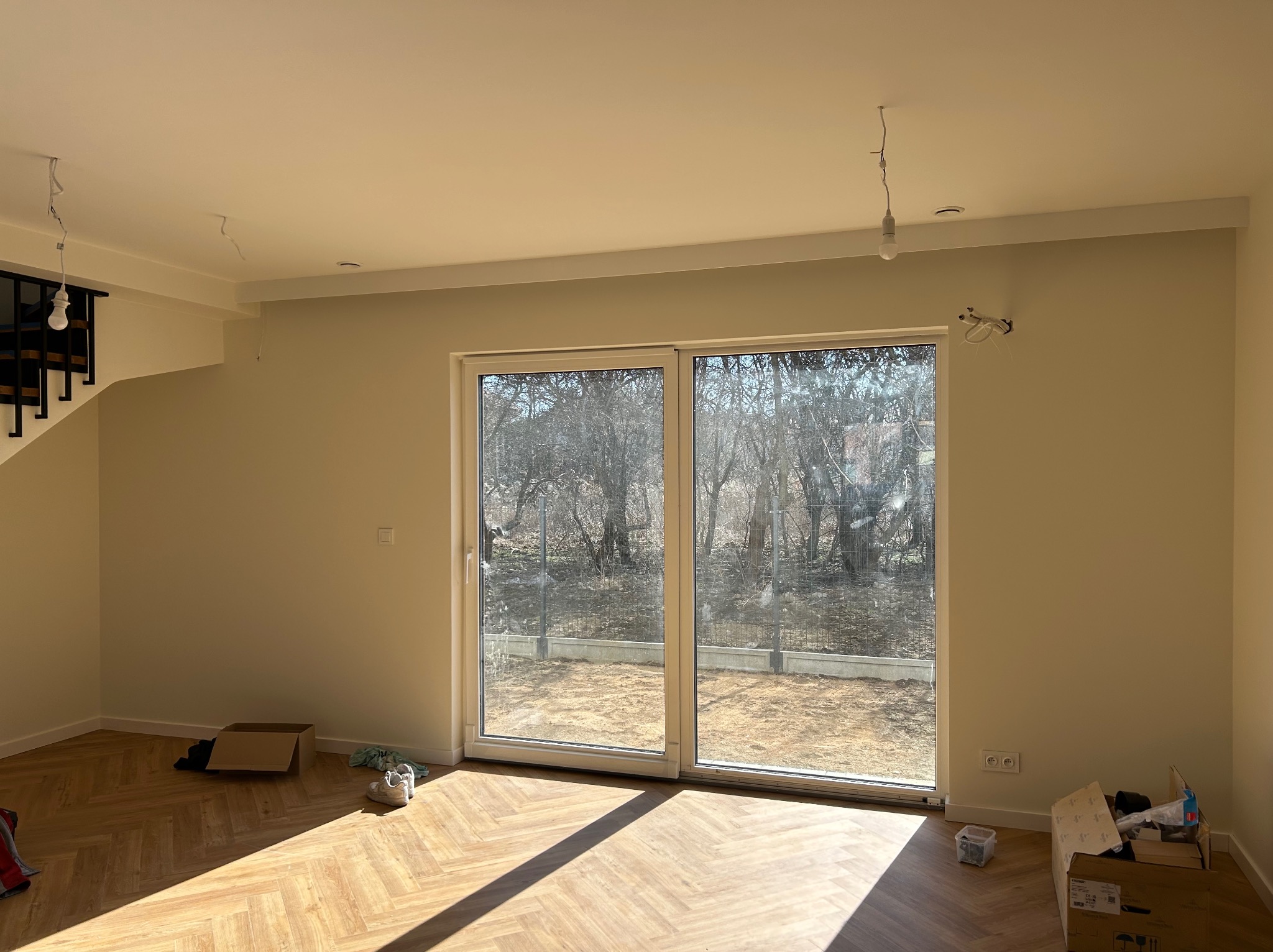 Jasne wnętrze z parkietem w jodełkę, duże okno balkonowe z widokiem na ogród. Ściany w odcieniach beżu, widoczne niedokończone instalacje elektryczne.