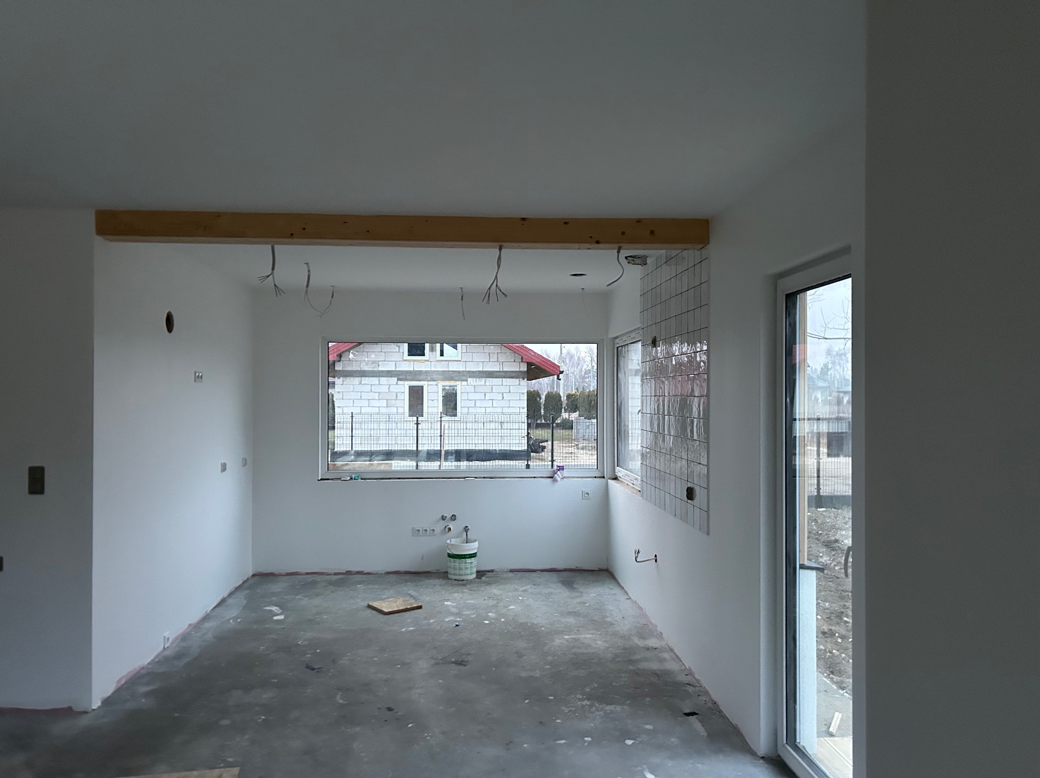 Surowe wnętrze w trakcie wykańczania: białe ściany, surowy beton na podłodze, widoczne instalacje elektryczne, fragment ułożonych płytek i drewniany element konstrukcyjny.