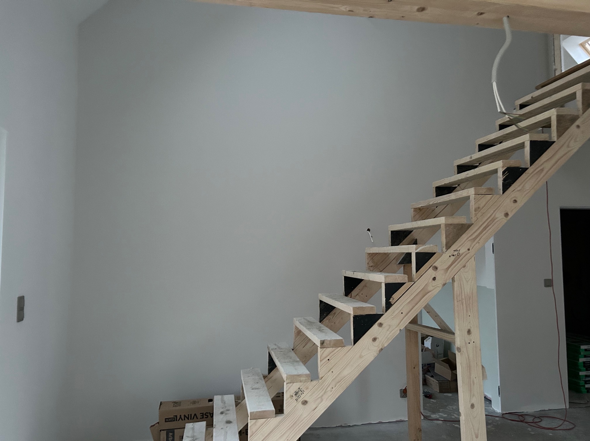 Surowe drewniane schody w jasnym wnętrzu, ściany przygotowane do malowania, widoczne elementy instalacji elektrycznej. Etap wykończenia wnętrza.