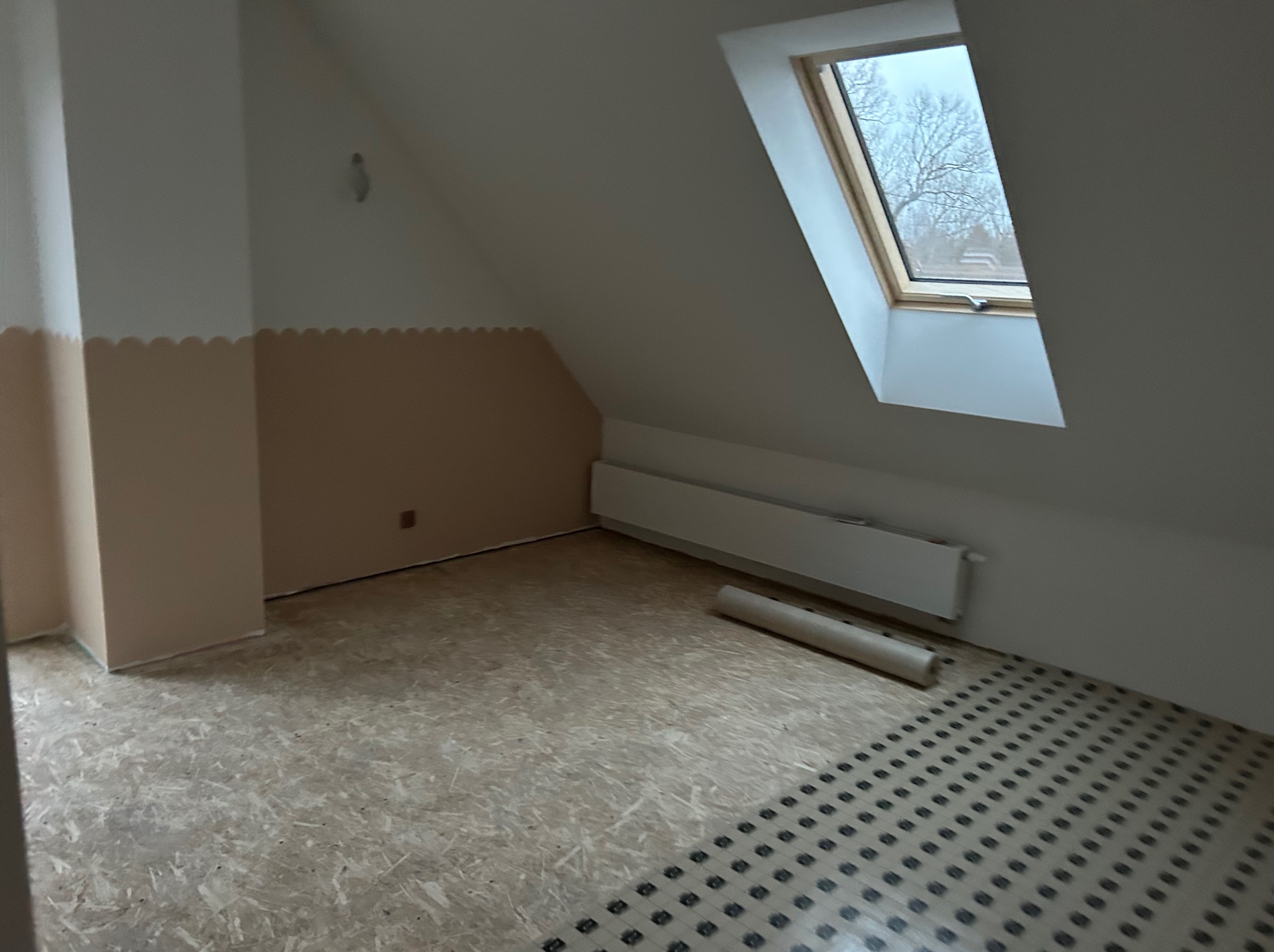 Poddasze po szpachlowaniu ścian i przygotowaniu podłogi pod ogrzewanie. Widoczne okno dachowe i grzejnik. Ściany pomalowane do połowy na beżowo.