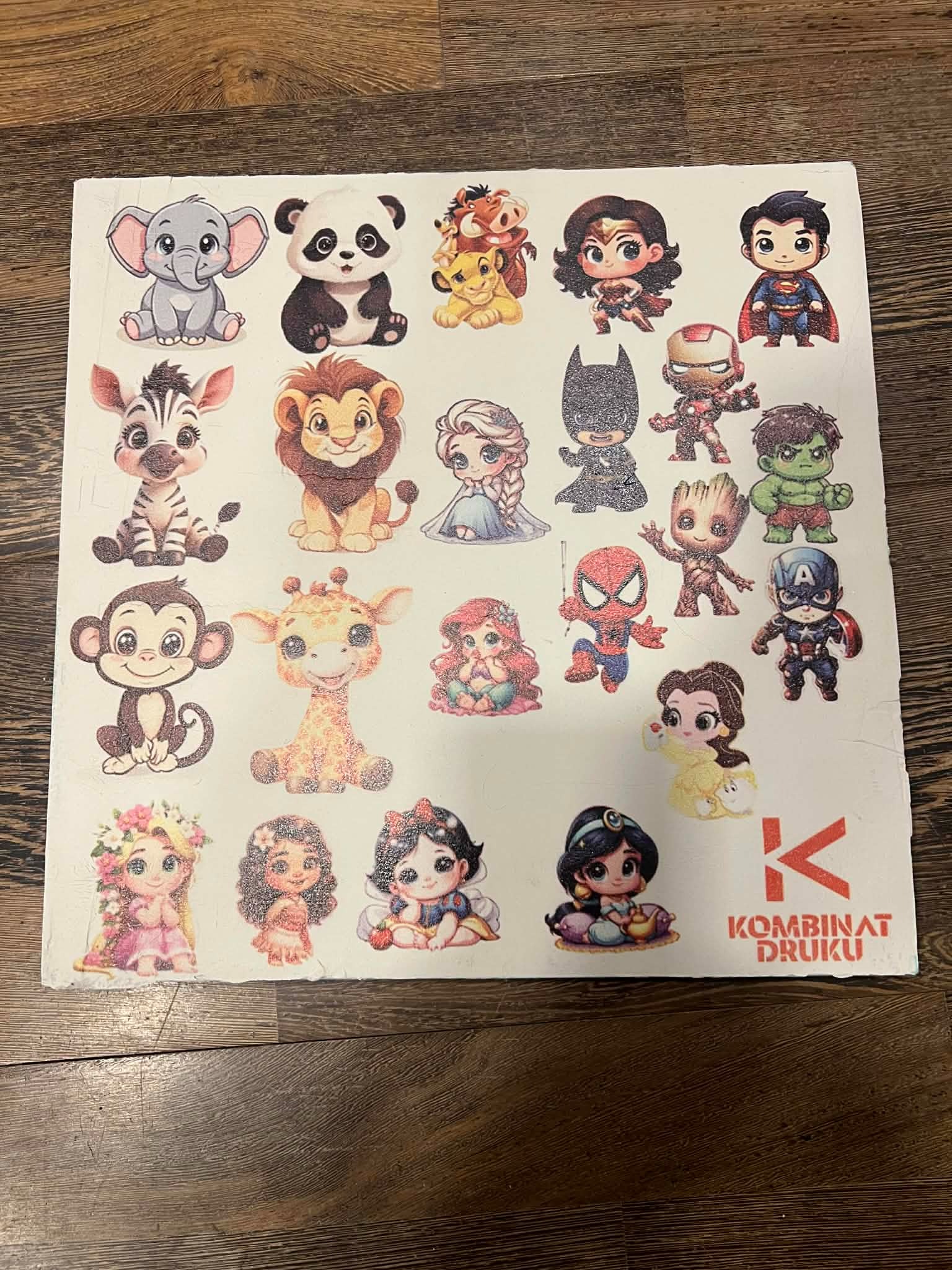 Arkusz naklejek z postaciami z bajek i superbohaterów, ułożony na drewnianym podłożu. Widoczne logo drukarni w prawym dolnym rogu.