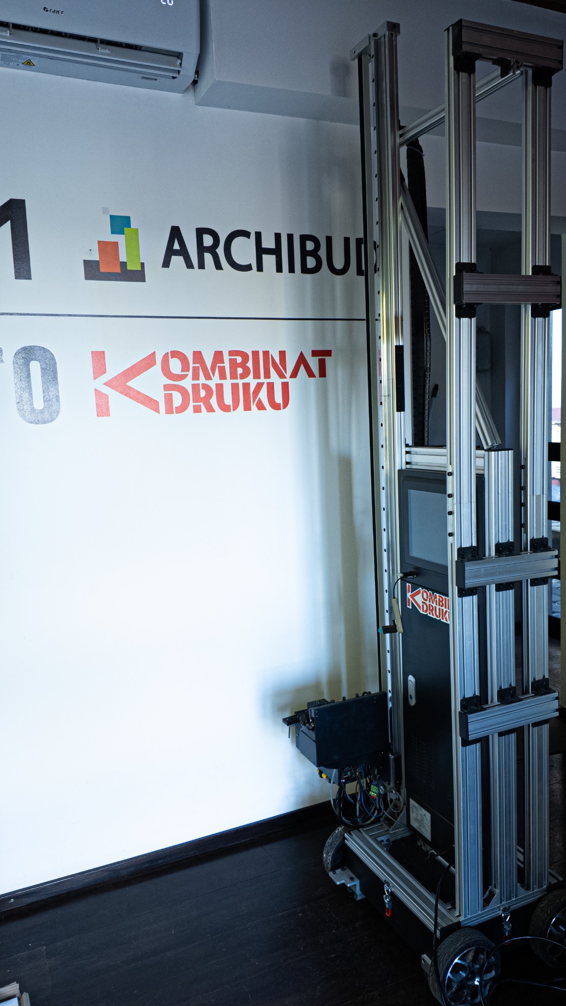 Drukarka pionowa do ścian z logo 'Kombinat Druku' na ścianie. Urządzenie na kółkach, z ekranem i aluminiową konstrukcją. Widoczny klimatyzator na górze.