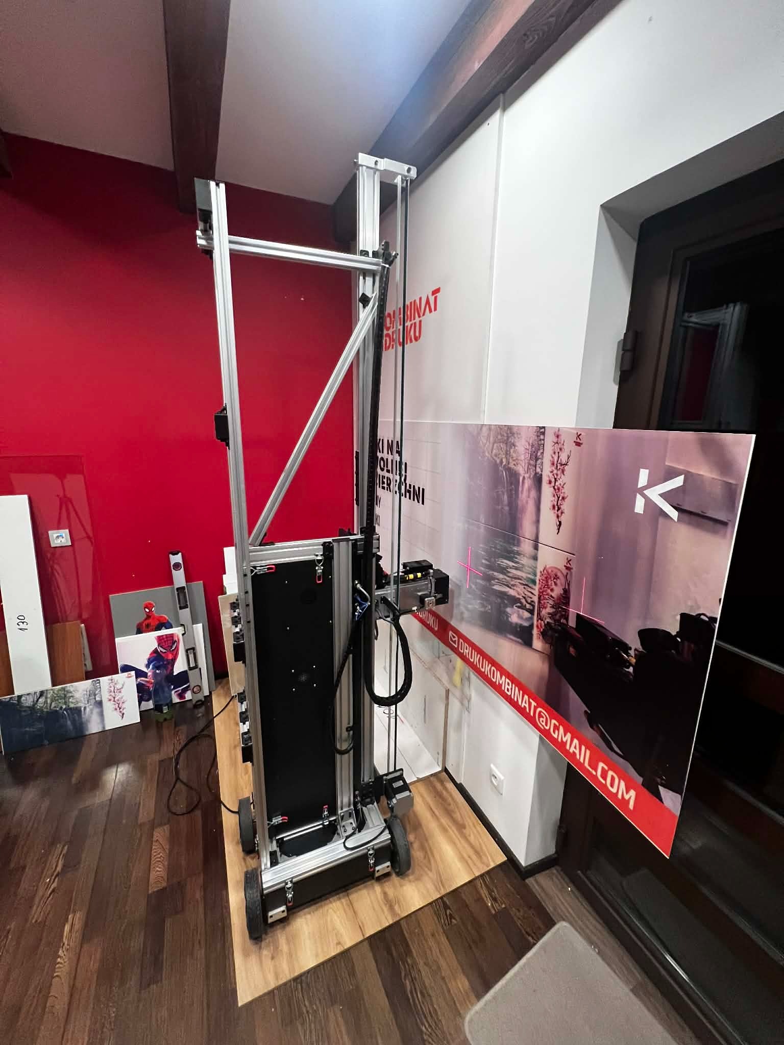 Wertykalna drukarka wielkoformatowa w trakcie druku plakatu z motywem krajobrazu i kart do gry. Widoczny adres e-mail drukukombinat@gmail.com.