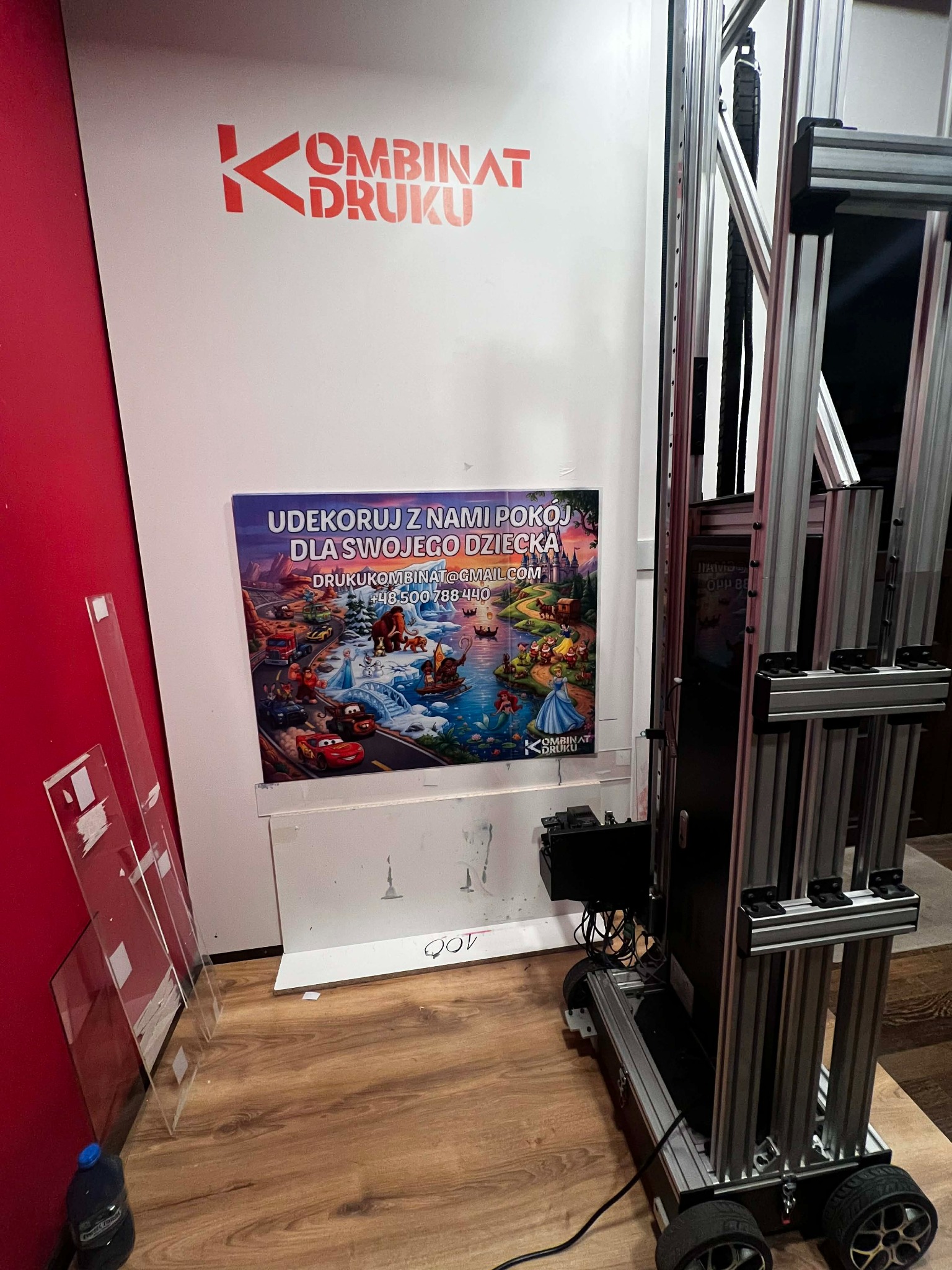 Drukarka wielkoformatowa w trakcie drukowania plakatu z motywem dziecięcym. Widoczne logo firmy 'Kombinat Druku'.