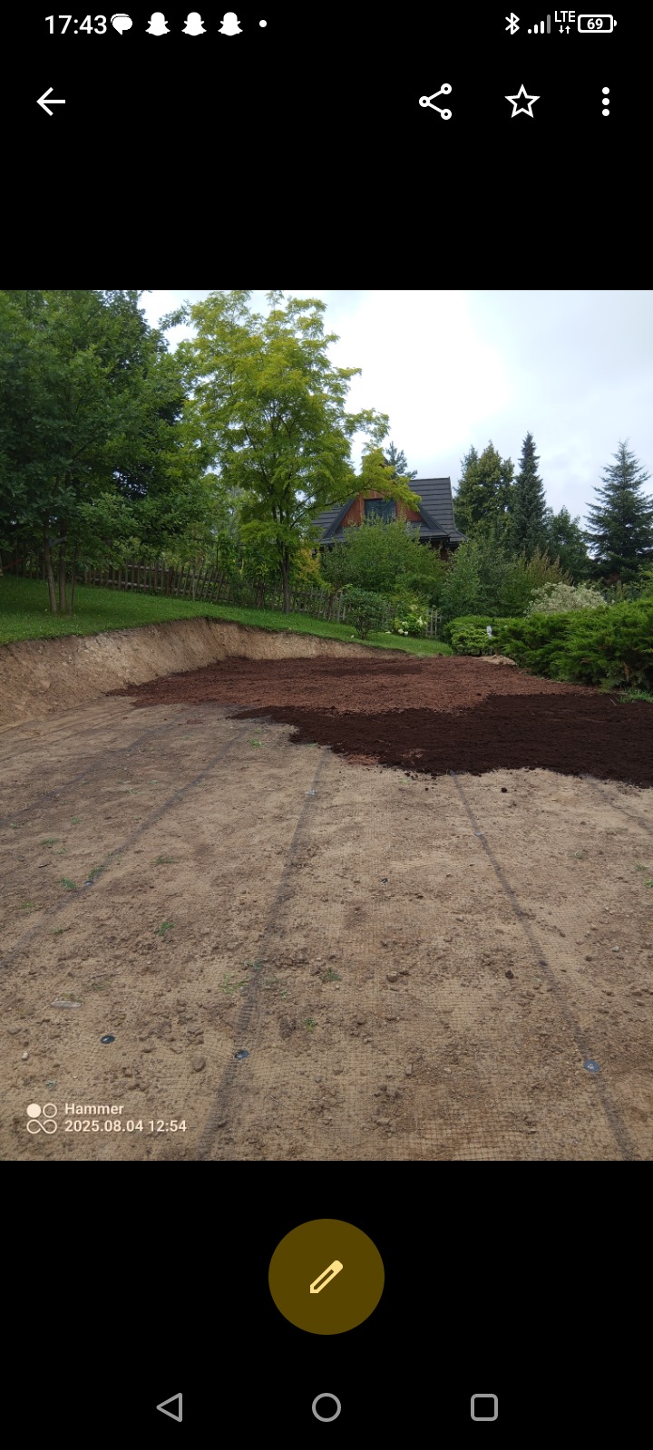Przygotowanie terenu pod trawnik: rozłożona siatka wzmacniająca i częściowo rozsypana warstwa żyznej ziemi na piaszczystym podłożu. W tle dom i zieleń.