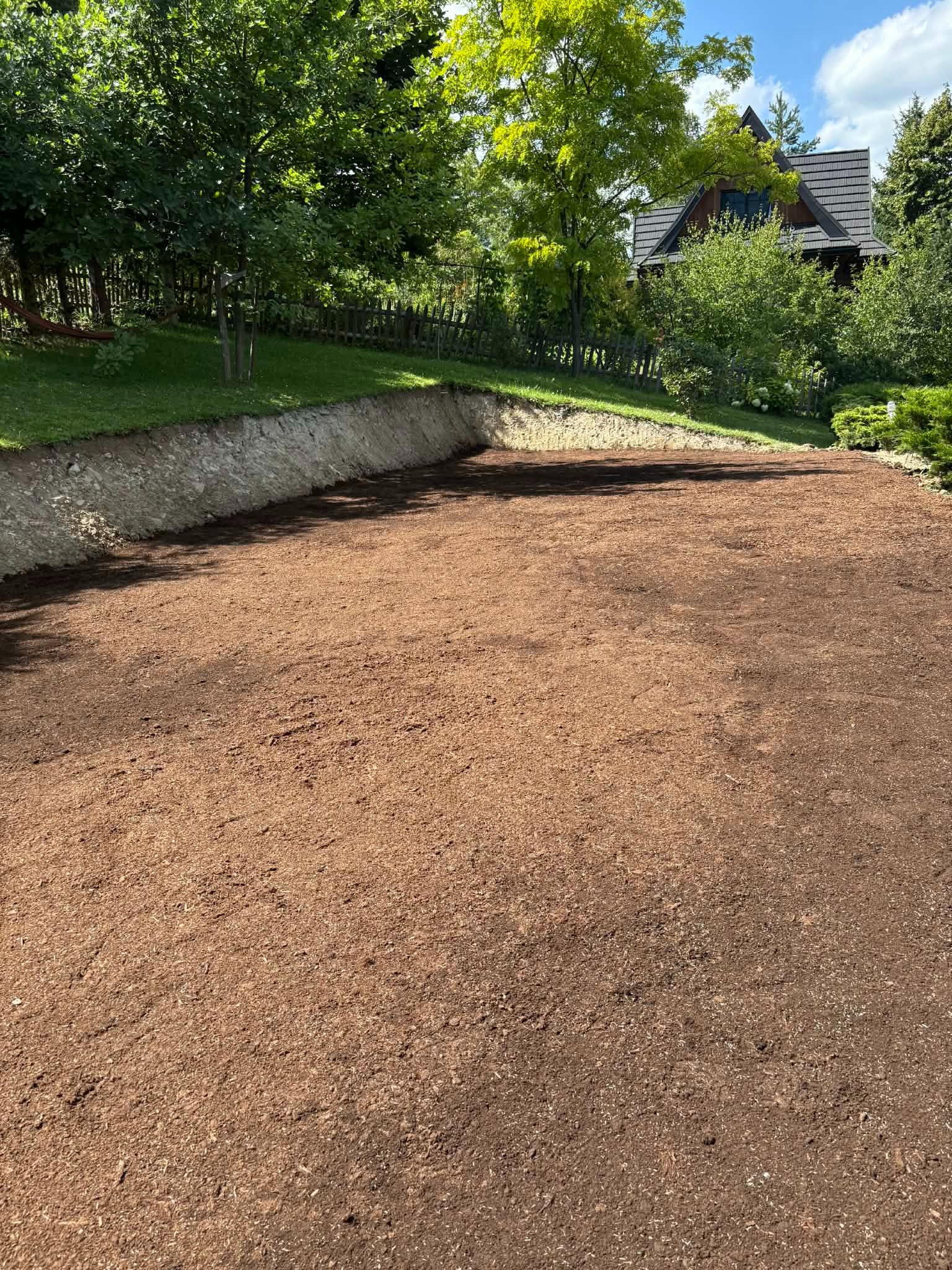 Przygotowany teren pod trawnik z widoczną warstwą ziemi urodzajnej, otoczony zielenią i domem w tle. Ujęcie z góry ukazuje równomierne rozłożenie podłoża.