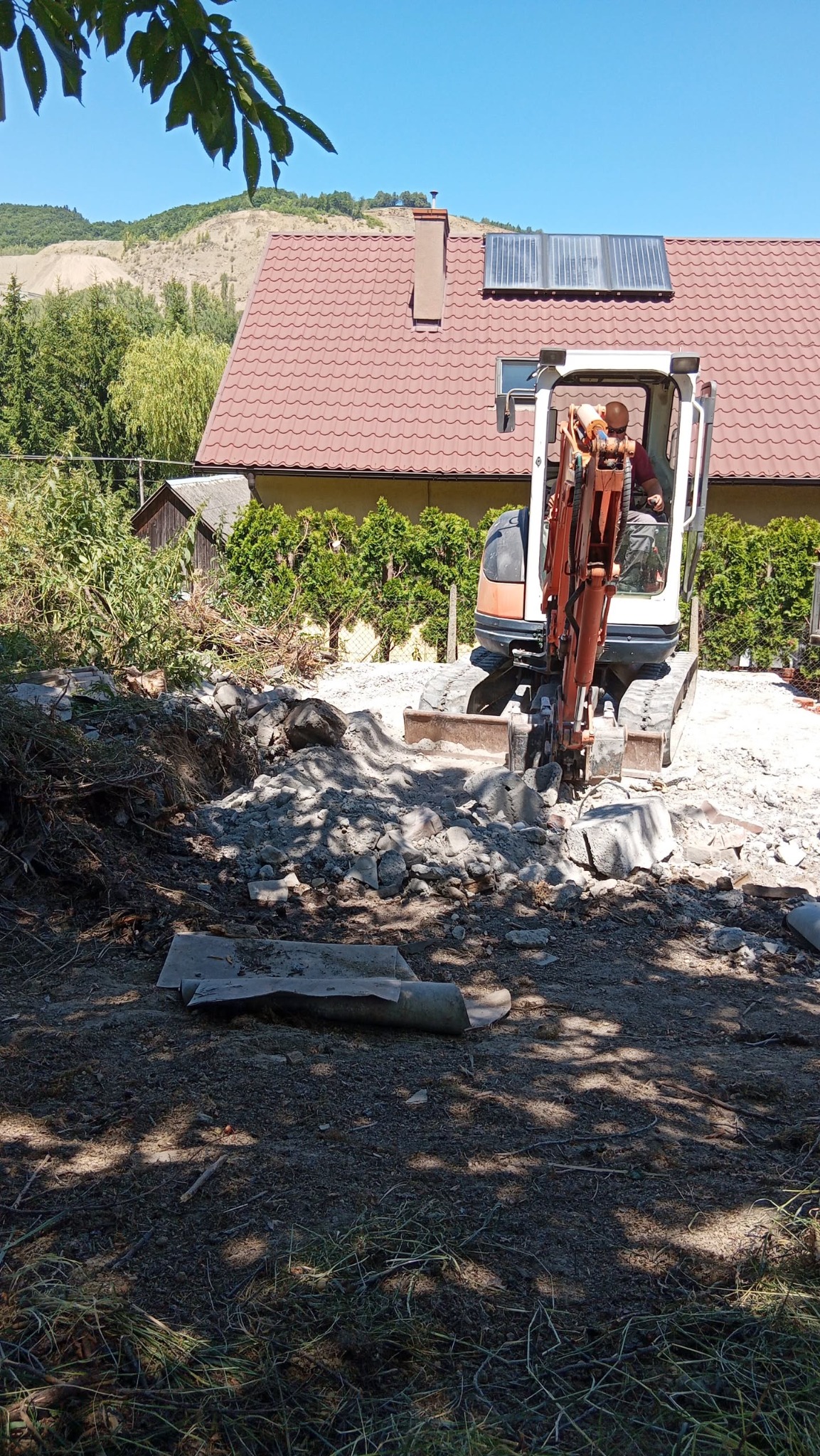 Mini koparka w akcji podczas prac ziemnych w Łososinie Dolnej. Widoczne gruz, ziemia i operator maszyny. W tle dom z czerwoną dachówką i zielone krzewy.