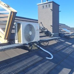 Monomon - Biała jednostka zewnętrzna klimatyzacji zamontowana na dachu z szarej dachówki, obok komina i okna dachowego. Widoczne przewody i metalowa konstrukcja wsporcza.