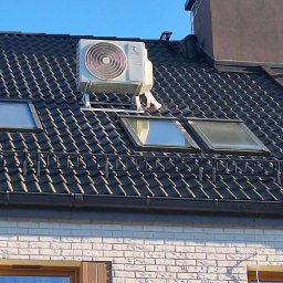 Monomon - Biała jednostka zewnętrzna klimatyzacji zamontowana na dachu domu z czarną dachówką i oknami dachowymi. Widoczny komin i fragment ściany z cegły.