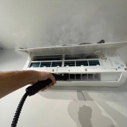 Monomon - Czyszczenie klimatyzacji parą wodną. Otwarta obudowa, widoczne filtry i dysza parowa w dłoni. Para wydobywa się z urządzenia. Białe ściany i sufit w tle.