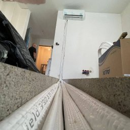 Monomon - Montaż klimatyzacji: widok z dołu na rury instalacyjne prowadzące do jednostki wewnętrznej na ścianie, z widocznymi elementami instalacji elektrycznej i kartonem AUX.