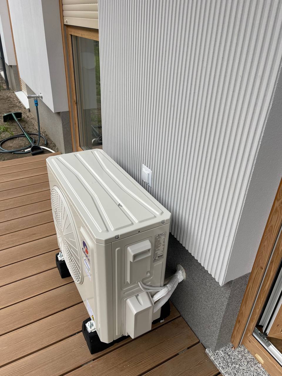 Biały klimatyzator zewnętrzny marki Gree, zamontowany na drewnianym tarasie przy budynku z elewacją w pionowe pasy. Widoczne przewody i czarne podkładki pod urządzeniem.