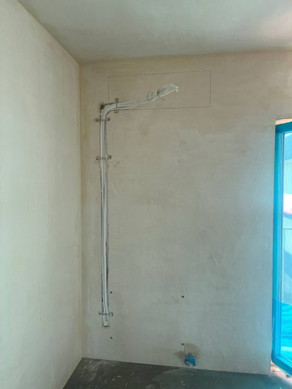 Białe rury instalacyjne, przymocowane do surowej ściany wewnątrz budynku, prowadzone pionowo i poziomo, z zaznaczonym prostokątem na ścianie.