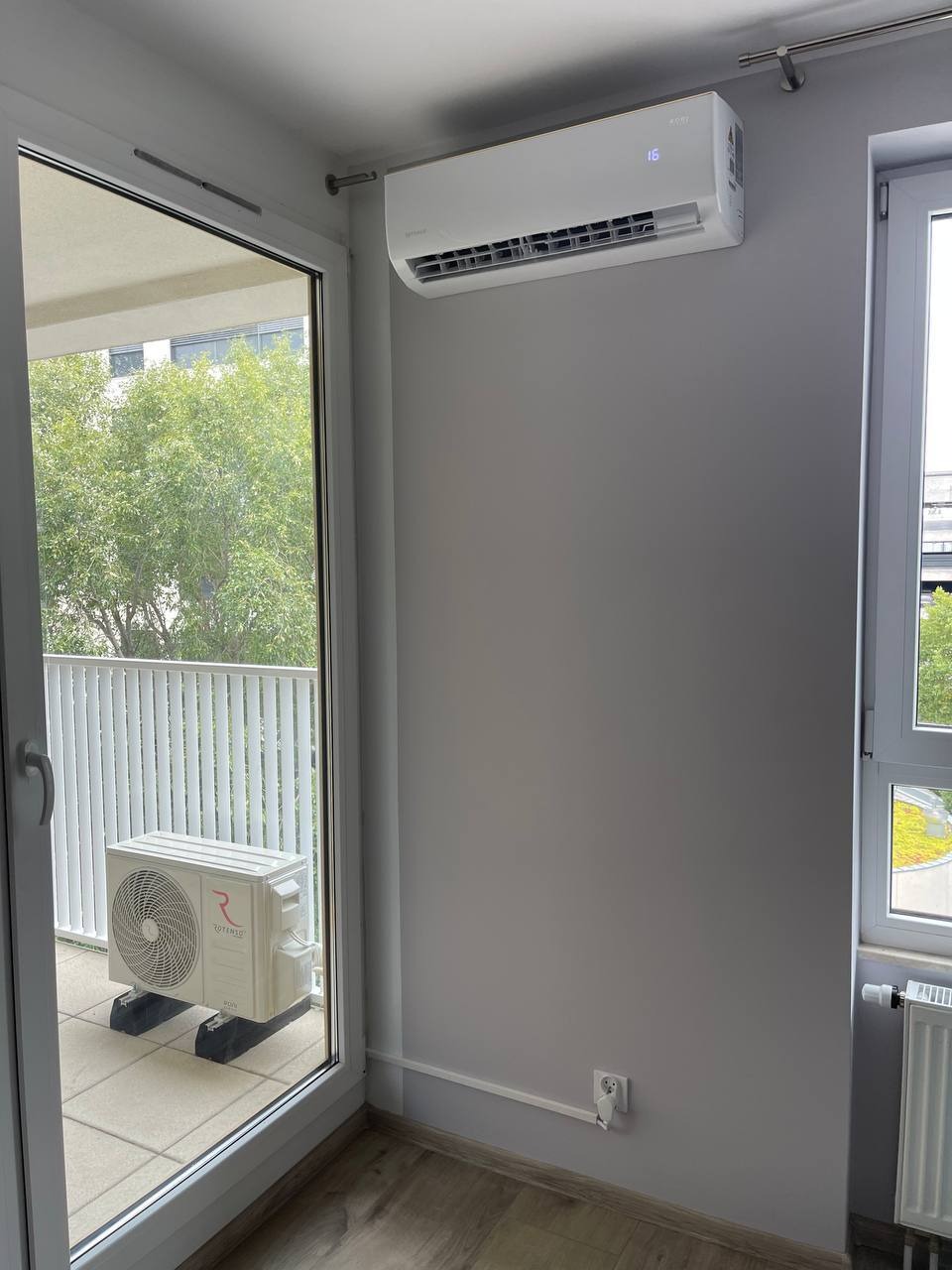 Biała klimatyzacja Rotenso zamontowana w jasnym pokoju z balkonem. Jednostka wewnętrzna na ścianie, zewnętrzna na balkonie. Widoczne okno i grzejnik.