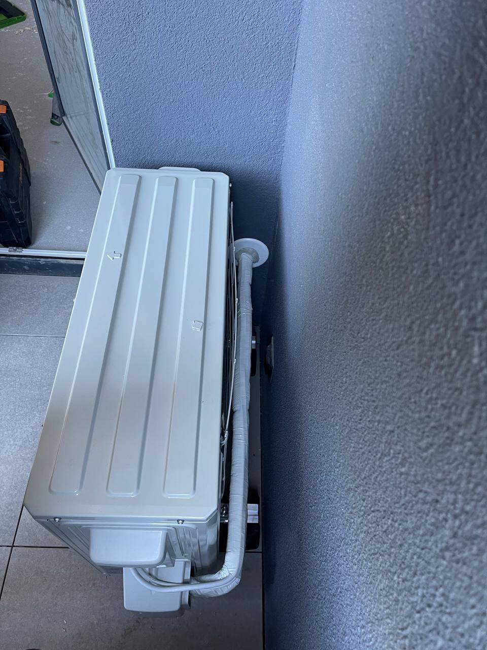 Zamontowana jednostka wewnętrzna klimatyzacji na szarej ścianie, widoczne rury odprowadzające skropliny, fragment podłogi z szarych płytek i skrzynka narzędziowa w tle.