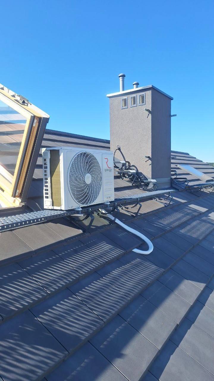 Biała jednostka zewnętrzna klimatyzacji zamontowana na dachu z szarej dachówki, obok komina i okna dachowego. Widoczne przewody i metalowa konstrukcja wsporcza.