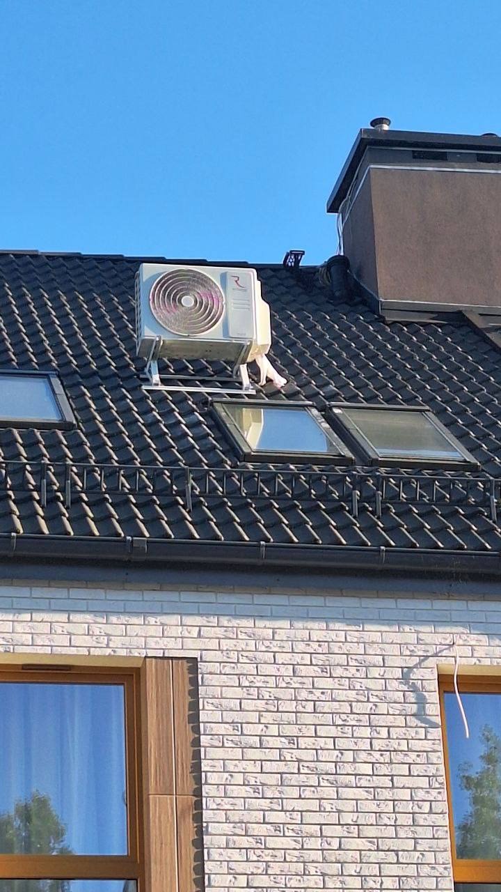 Biała jednostka zewnętrzna klimatyzacji zamontowana na dachu domu z czarną dachówką i oknami dachowymi. Widoczny komin i fragment ściany z cegły.