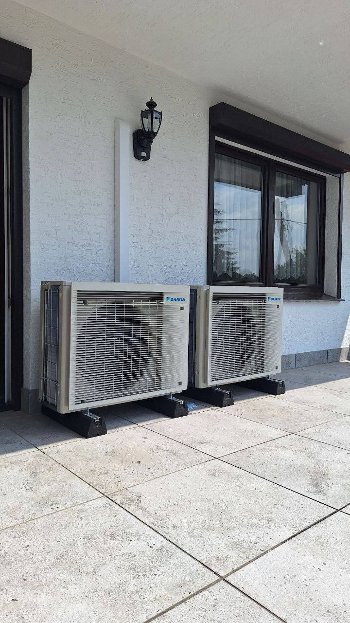 Dwa klimatyzatory Daikin na zewnątrz budynku z oknem i lampą. Urządzenia stoją na czarnych podstawach, na jasnej, kwadratowej nawierzchni.