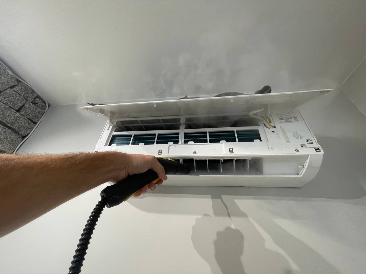 Czyszczenie klimatyzacji parą wodną. Otwarta obudowa, widoczne filtry i dysza parowa w dłoni. Para wydobywa się z urządzenia. Białe ściany i sufit w tle.