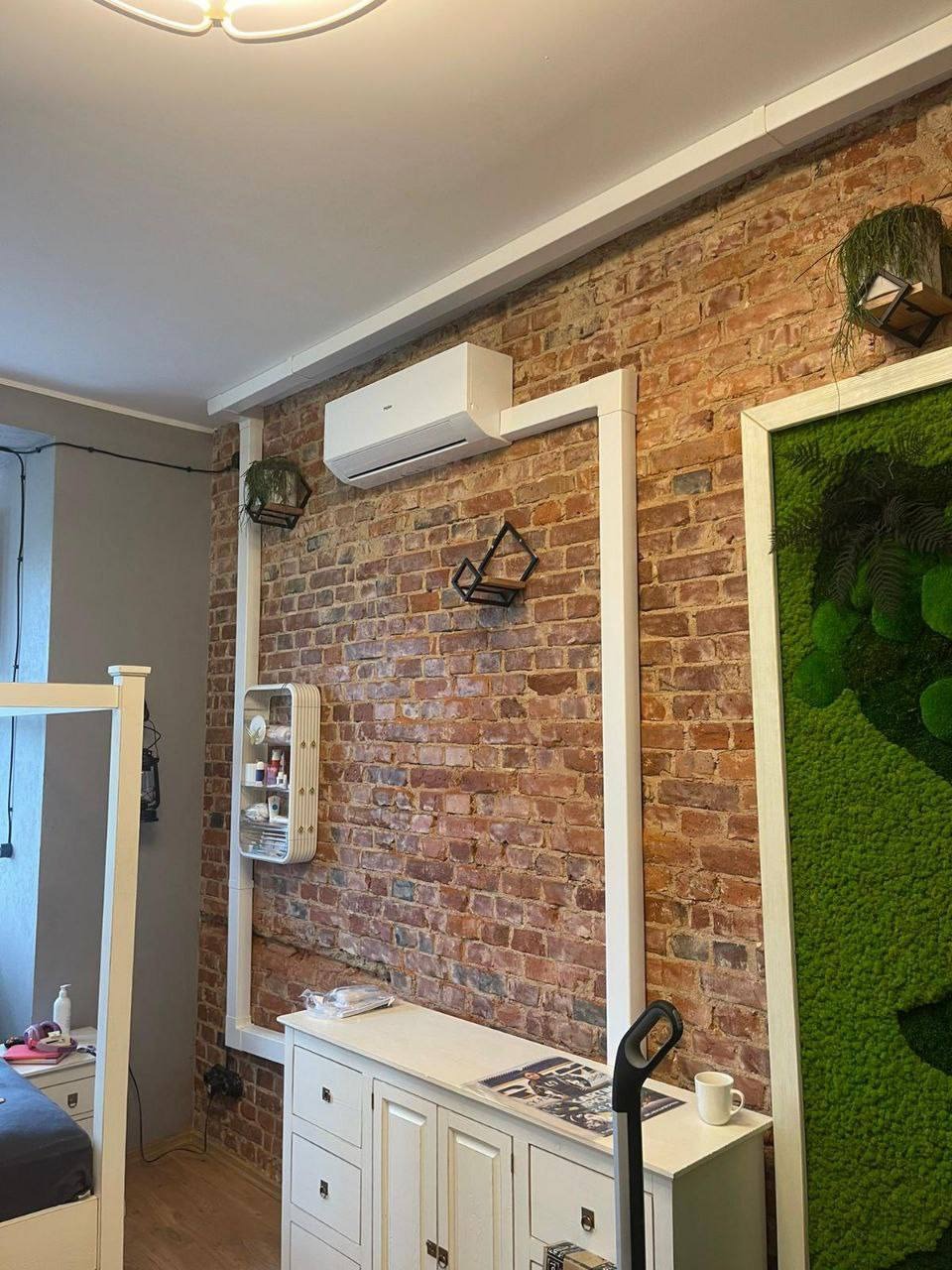 Biały klimatyzator na ceglanej ścianie w pokoju z dekoracjami roślinnymi i białymi meblami. Widoczne białe rury instalacyjne.