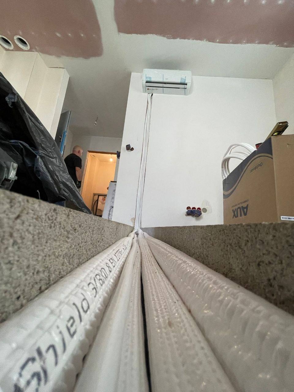 Montaż klimatyzacji: widok z dołu na rury instalacyjne prowadzące do jednostki wewnętrznej na ścianie, z widocznymi elementami instalacji elektrycznej i kartonem AUX.