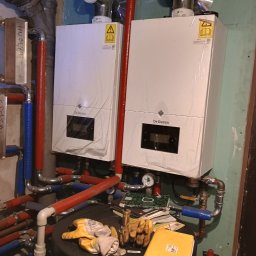 Hydraulik Przemek - Dwa białe, nowe piece gazowe De Dietrich zamontowane na ścianie, z widocznymi rurami instalacji. Na zbiorniku leżą rękawice robocze i dokumentacja.