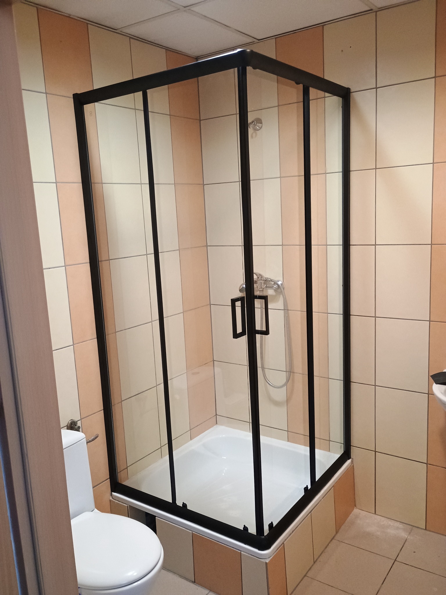 Kabina prysznicowa z czarną ramą w łazience z beżowo-pomarańczowymi płytkami. Widoczny fragment toalety i umywalki. Nowoczesny design.