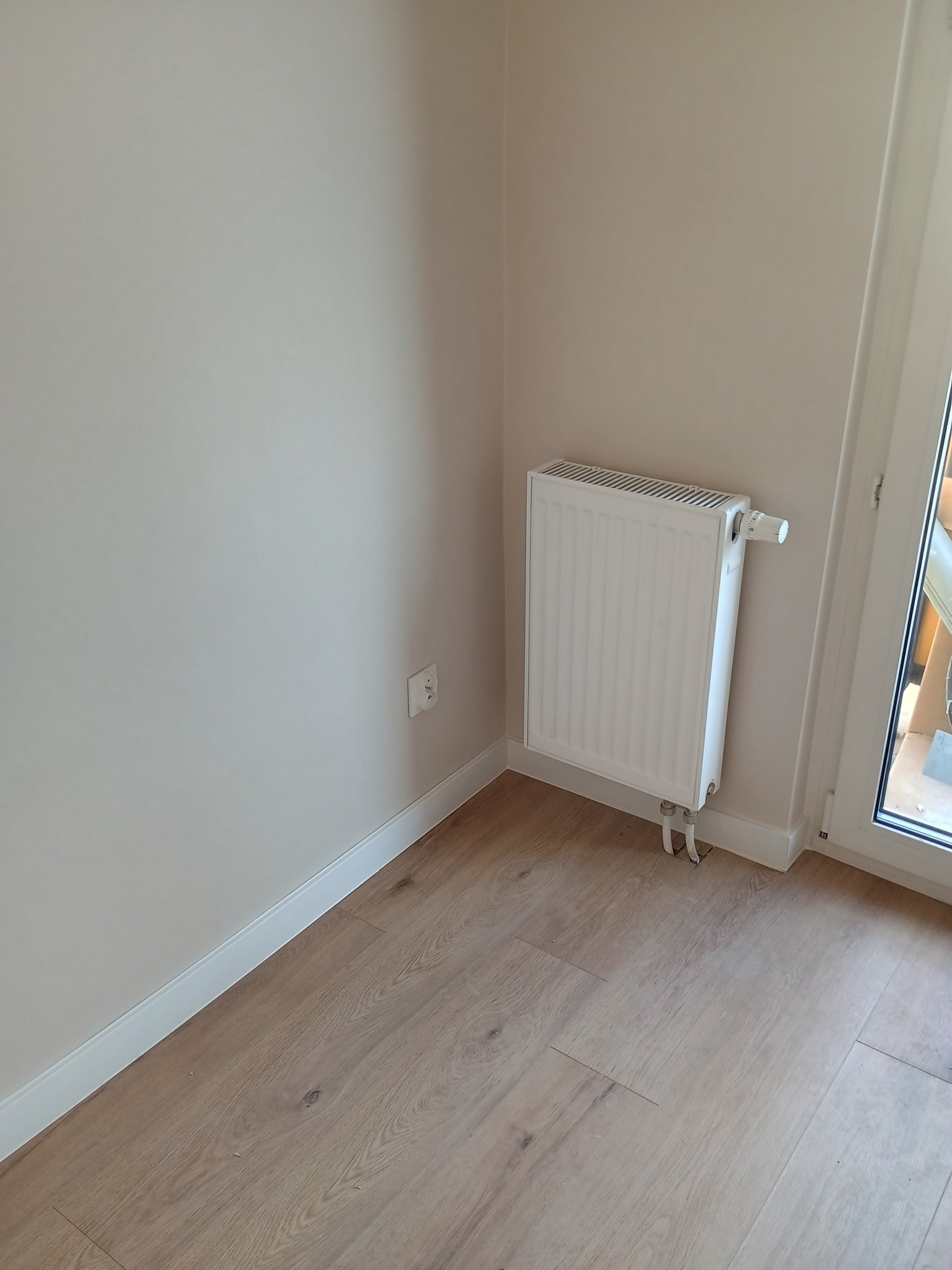 Biały grzejnik panelowy zamontowany przy ścianie w Miedzianej Górze. Widoczne drewniane panele podłogowe i listwy przypodłogowe. Ujęcie z niskiego kąta.