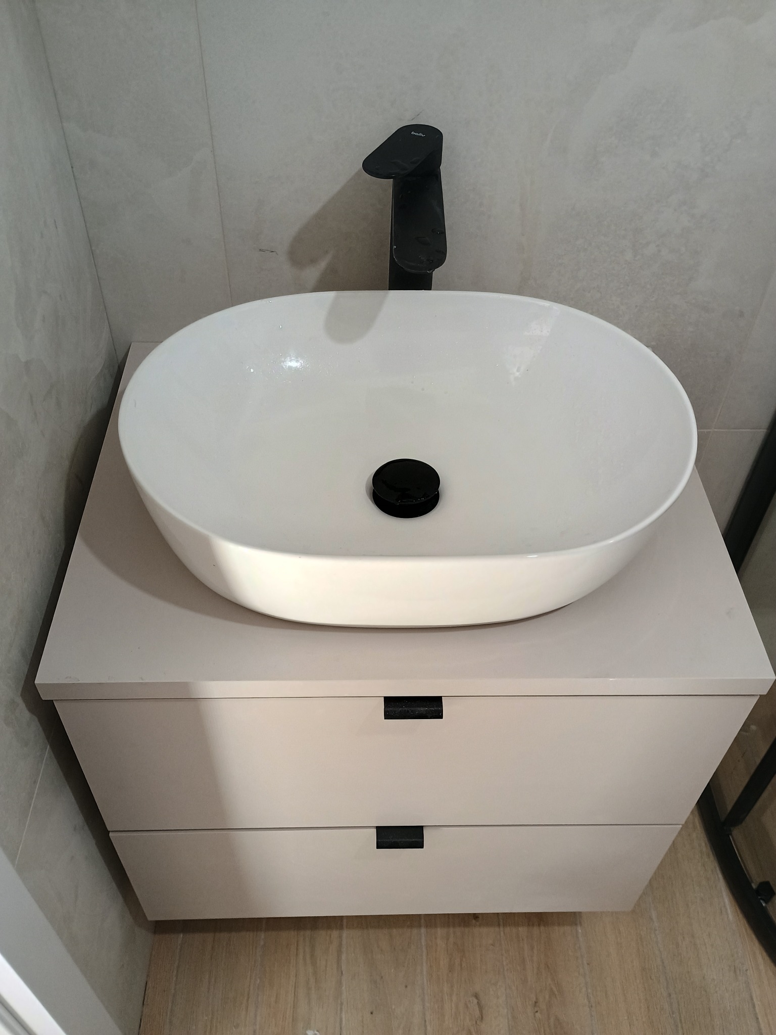 Biała, owalna umywalka nablatowa z czarną armaturą na szafce z dwiema szufladami. Minimalistyczny design, jasne płytki w tle, drewnopodobna podłoga.