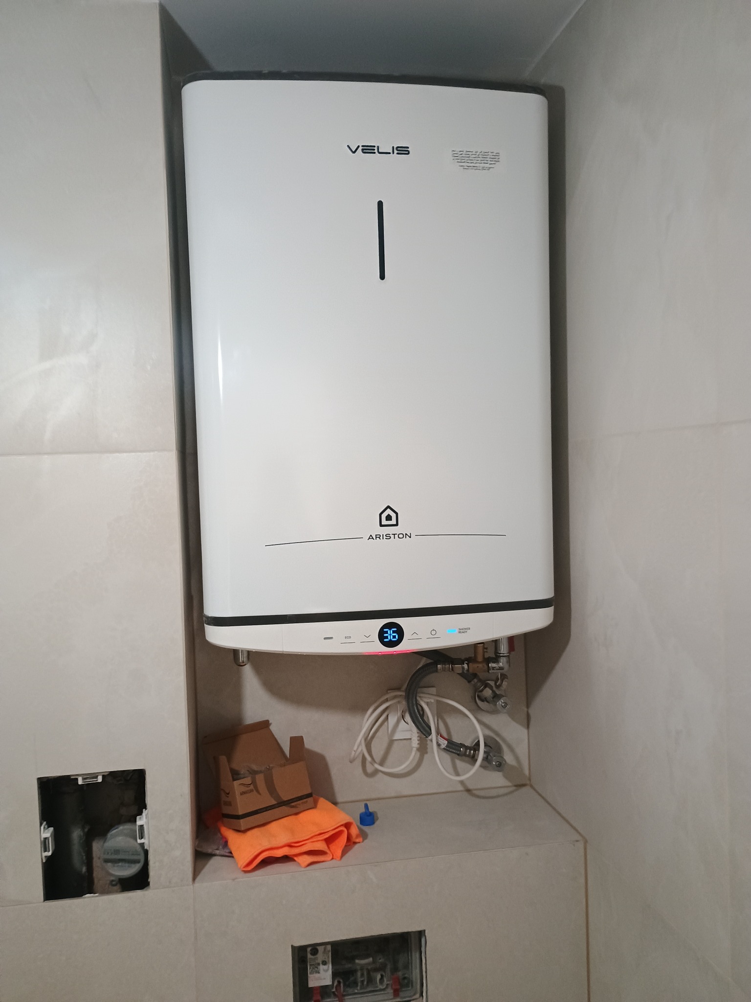 Biały, wiszący bojler Ariston Velis w łazience, z widocznymi przyłączami i fragmentem instalacji. W tle jasne płytki. Widoczny otwór rewizyjny i akcesoria montażowe.