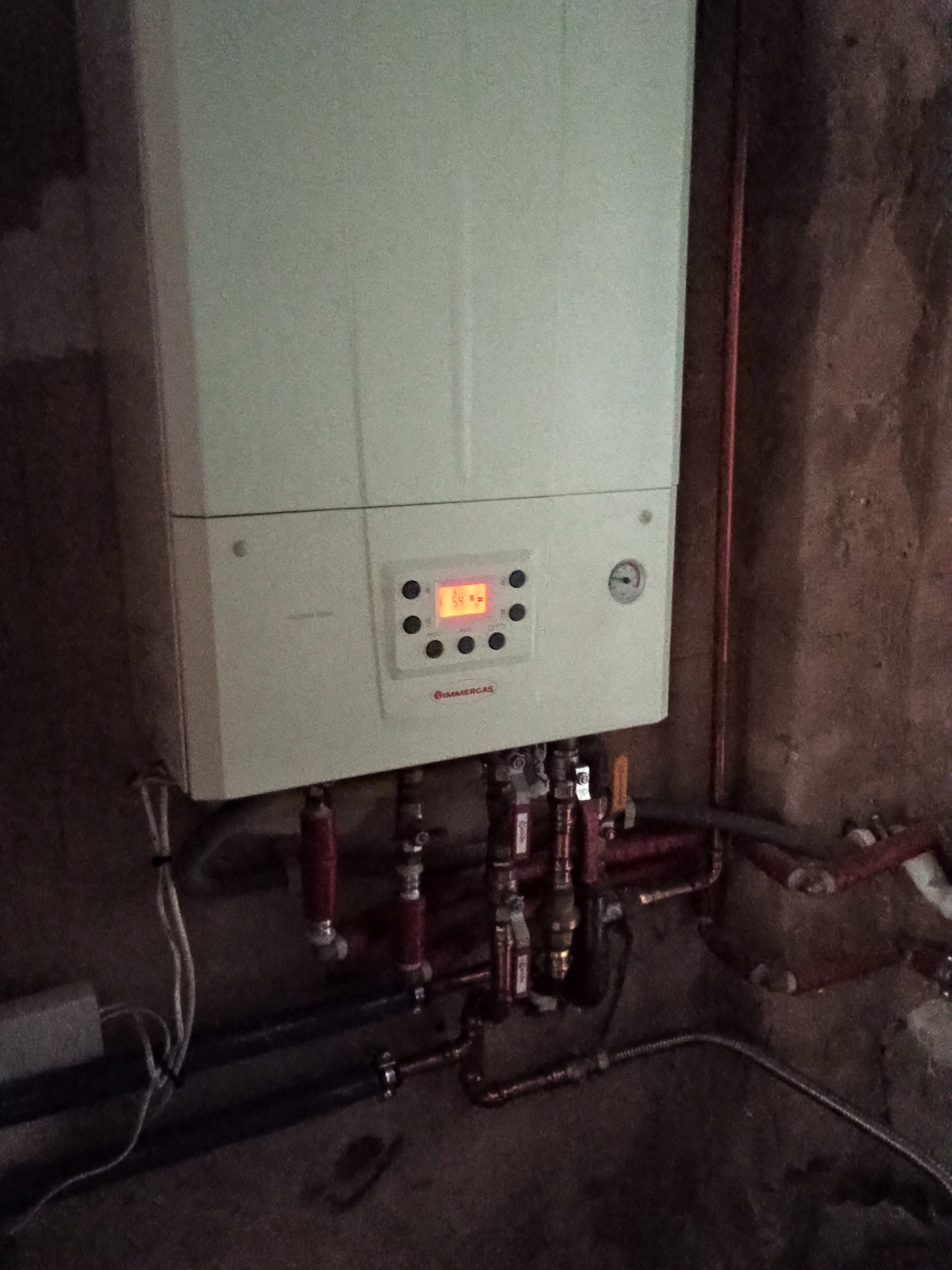 Zamontowany, biały piec gazowy marki Immergas z widocznymi rurami miedzianymi i zaworami, na tle ściany o surowej fakturze. Widoczny wyświetlacz z temperaturą.