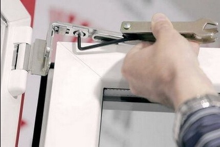 Regulacja zawiasu okiennego z użyciem klucza imbusowego i specjalistycznego narzędzia. Białe okno PCV, srebrny zawias, dłoń w zegarku. Detal.