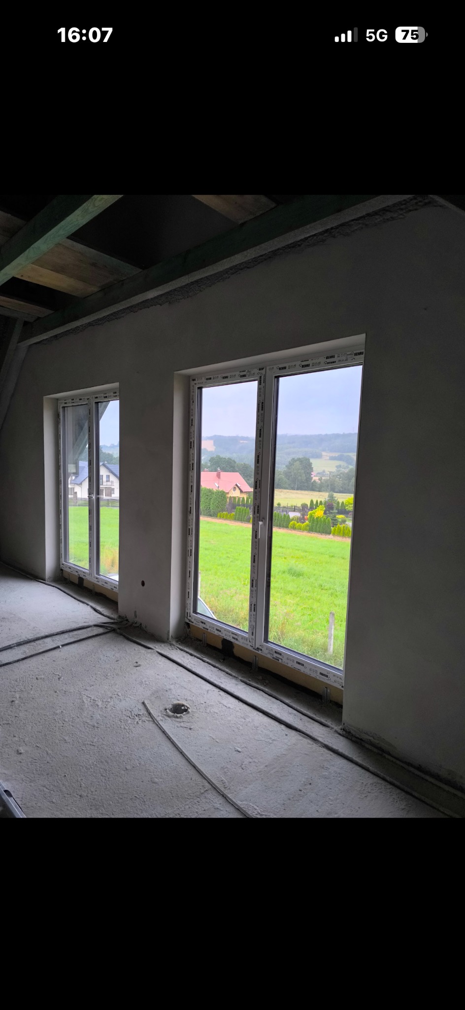 Wnętrze w stanie surowym z dwoma oknami z widokiem na zielony krajobraz. Ściany pokryte świeżym tynkiem, widoczne elementy instalacji na podłodze. Drewniana konstrukcja dachu.