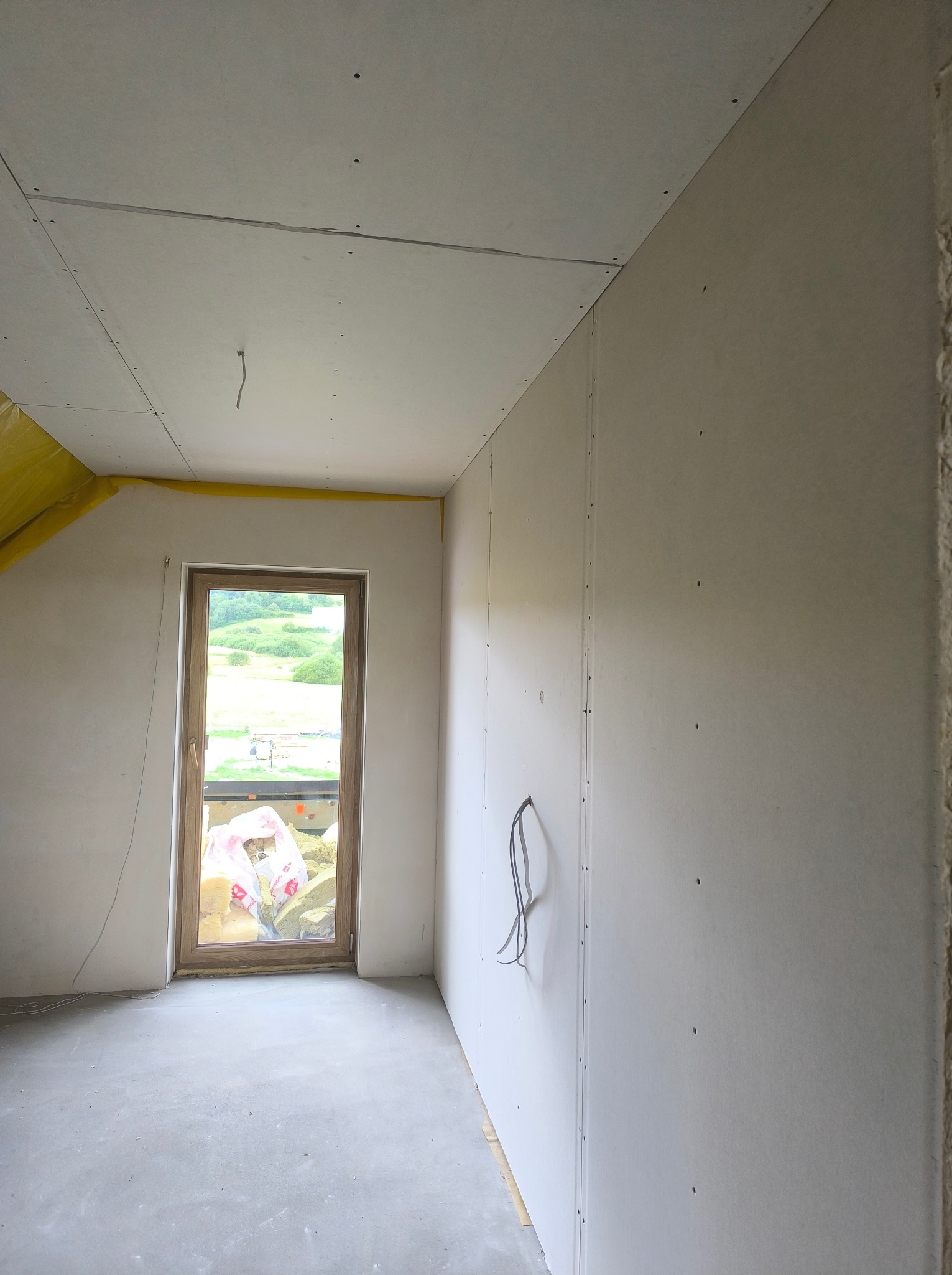 Poddasze z oknem po montażu płyt kartonowo-gipsowych na ścianach i suficie. Widoczne kable elektryczne i ocieplenie w narożniku. Surowy stan wykończenia wnętrza.
