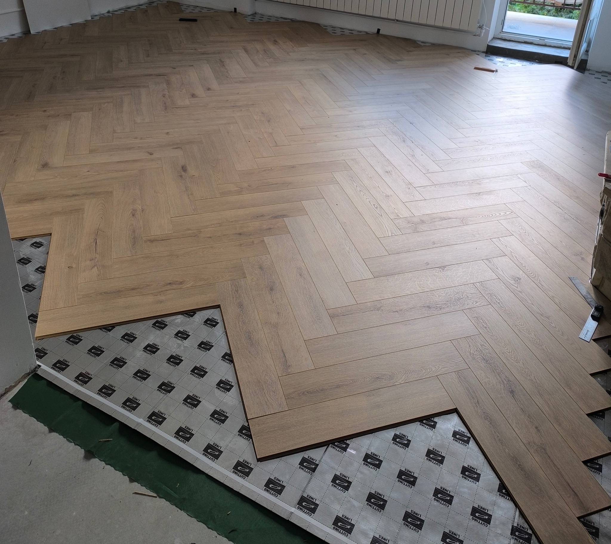 Układanie parkietu w jodełkę. Częściowo ułożona podłoga z jasnego drewna, widoczna podkładka pod panele. W tle okno i grzejnik.