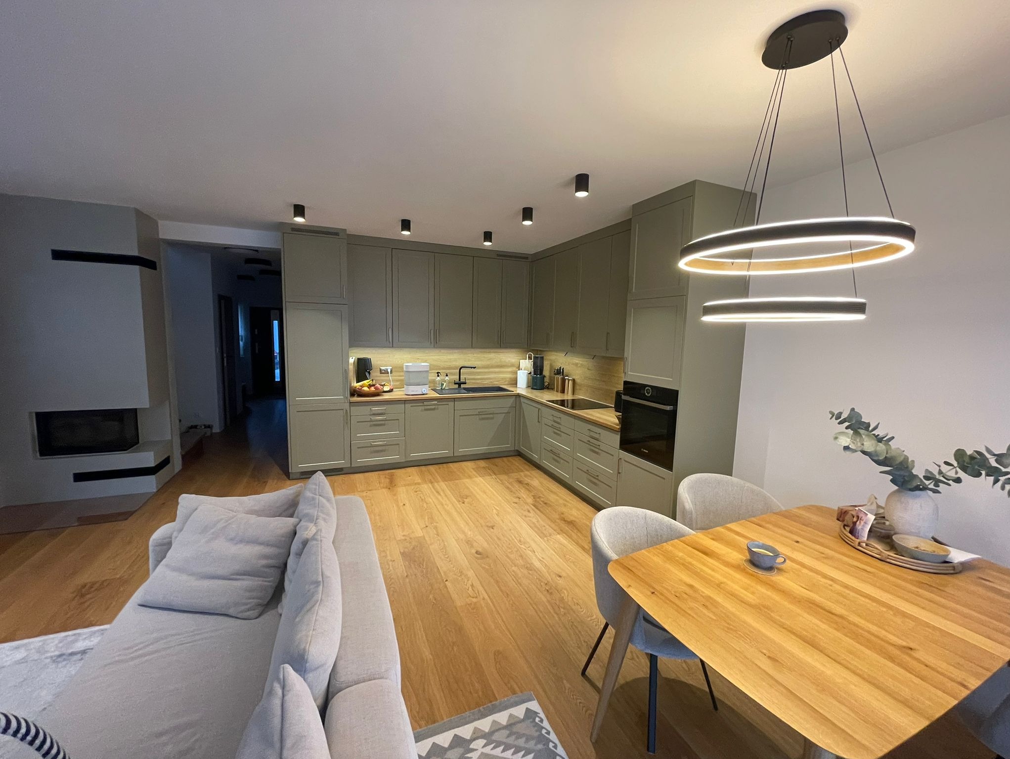 Nowoczesna kuchnia w odcieniach szarości z drewnianym blatem i podłogą. Nad stołem wiszą designerskie, okrągłe lampy. Widoczny fragment salonu z szarą sofą.