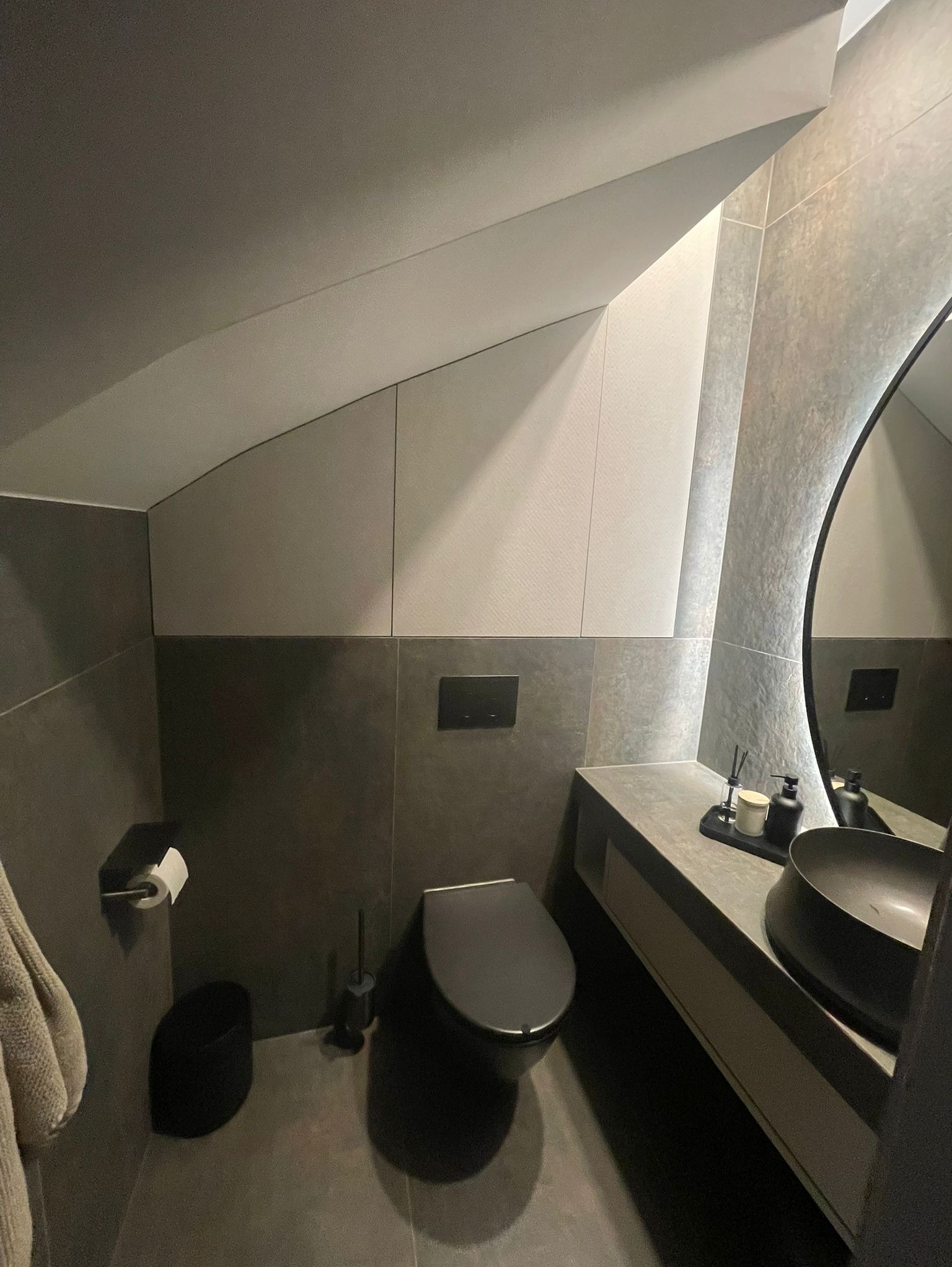 Nowoczesna łazienka na poddaszu w szarościach: owalne lustro z podświetleniem, umywalka nablatowa, wisząca toaleta i geometryczne wykończenie ścian.