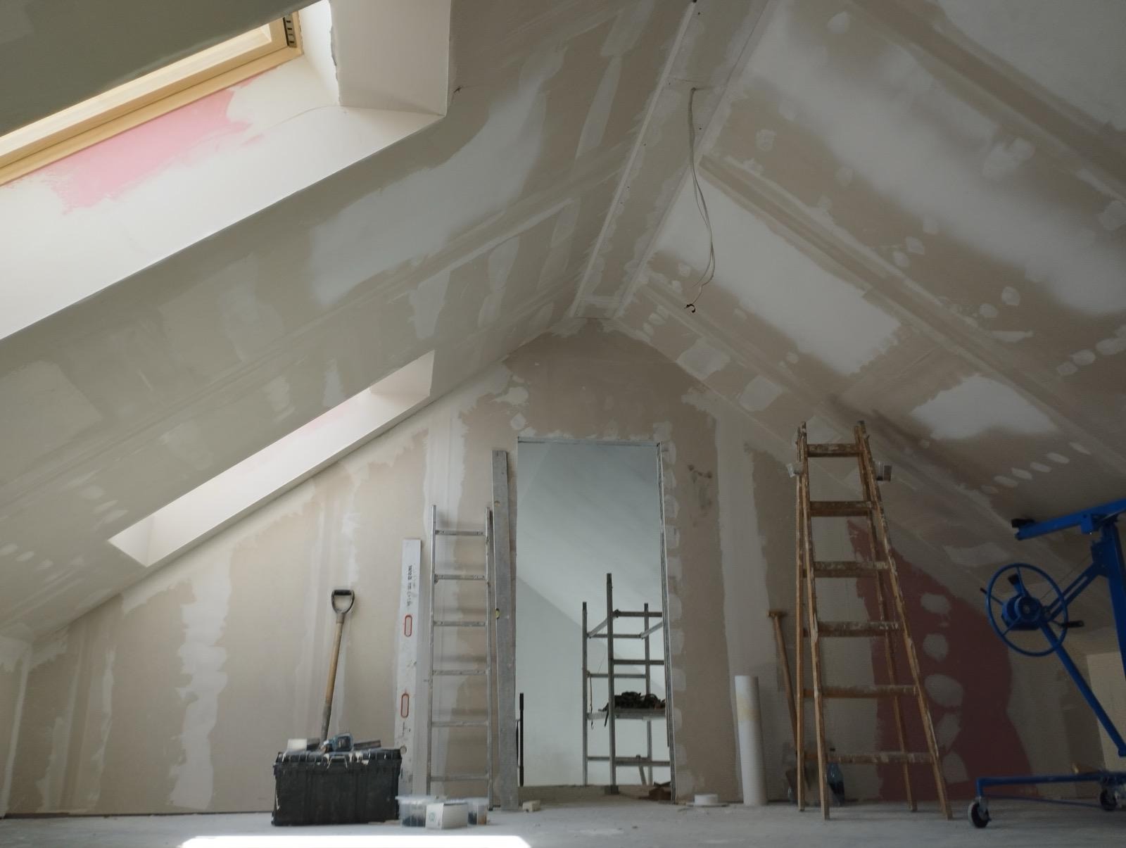 Poddasze w trakcie szpachlowania, widoczne okna dachowe, drabiny, narzędzia i podnośnik do płyt. Ściany i sufit pokryte szpachlą, przygotowane do dalszych prac wykończeniowych.