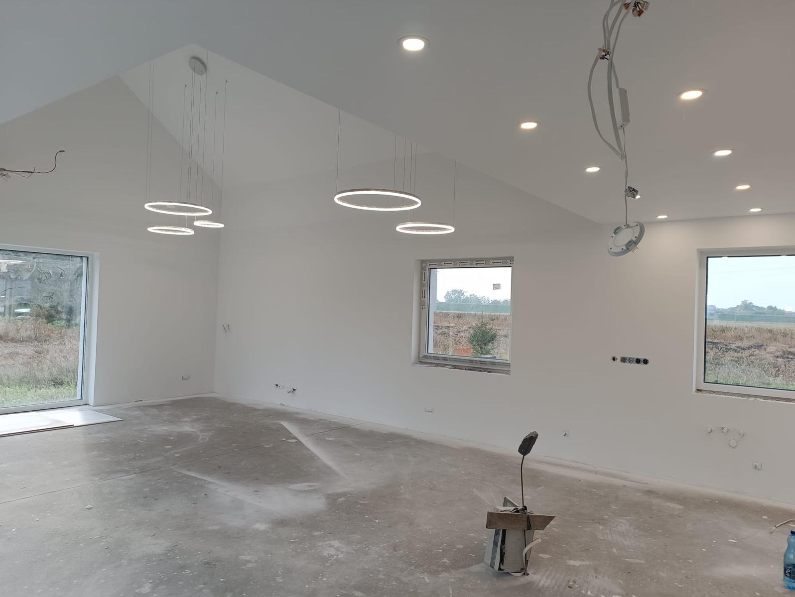 Jasne, przestronne wnętrze w trakcie wykańczania: białe ściany, surowa podłoga, okna z widokiem na zewnątrz i nowoczesne, okrągłe lampy wiszące. Widoczne niedokończone instalacje.