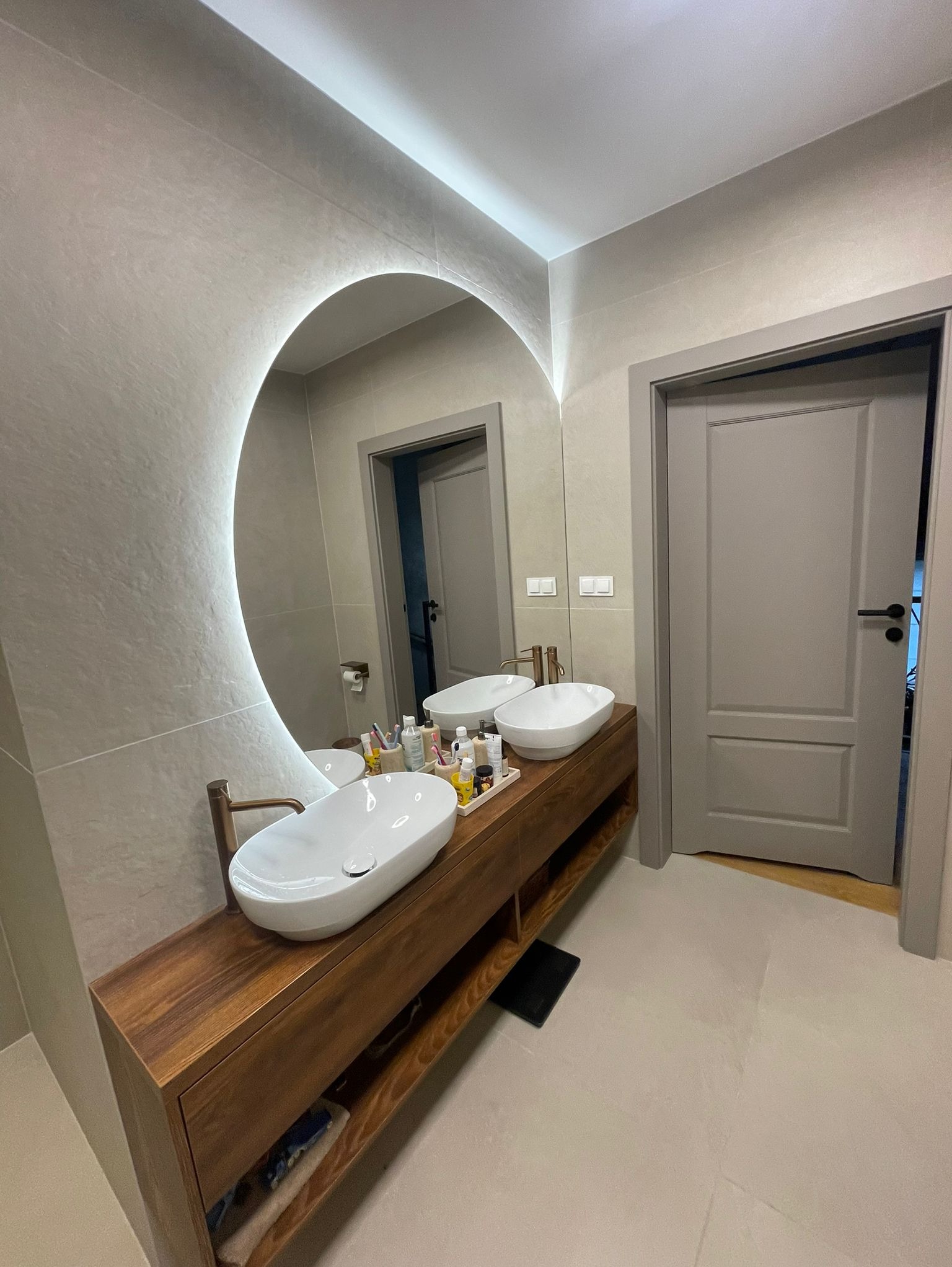 Nowoczesna łazienka z owalnym lustrem podświetlanym LED, drewnianą szafką pod umywalki i szarą armaturą. Minimalistyczny design, jasne płytki i drzwi w odcieniach szarości.