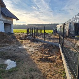 GABRO OGRODZENIA Bartosz Gabryszewski - Montaż ogrodzenia panelowego na fundamencie betonowym przy budowie domu. Widoczny fragment ogrodzenia, teren budowy, osoba i samochód dostawczy w tle.