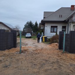 GABRO OGRODZENIA Bartosz Gabryszewski - Wjazd na posesję z częściowo zamontowaną bramą z betonowych bloczków. Widoczne słupki i przygotowany teren. W tle dom i samochód.