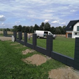 GABRO OGRODZENIA Bartosz Gabryszewski - Wznoszenie nowoczesnego, grafitowego ogrodzenia modułowego na trawiastym terenie. Widoczny fragment ogrodzenia, pracownik i biały samochód dostawczy w tle.