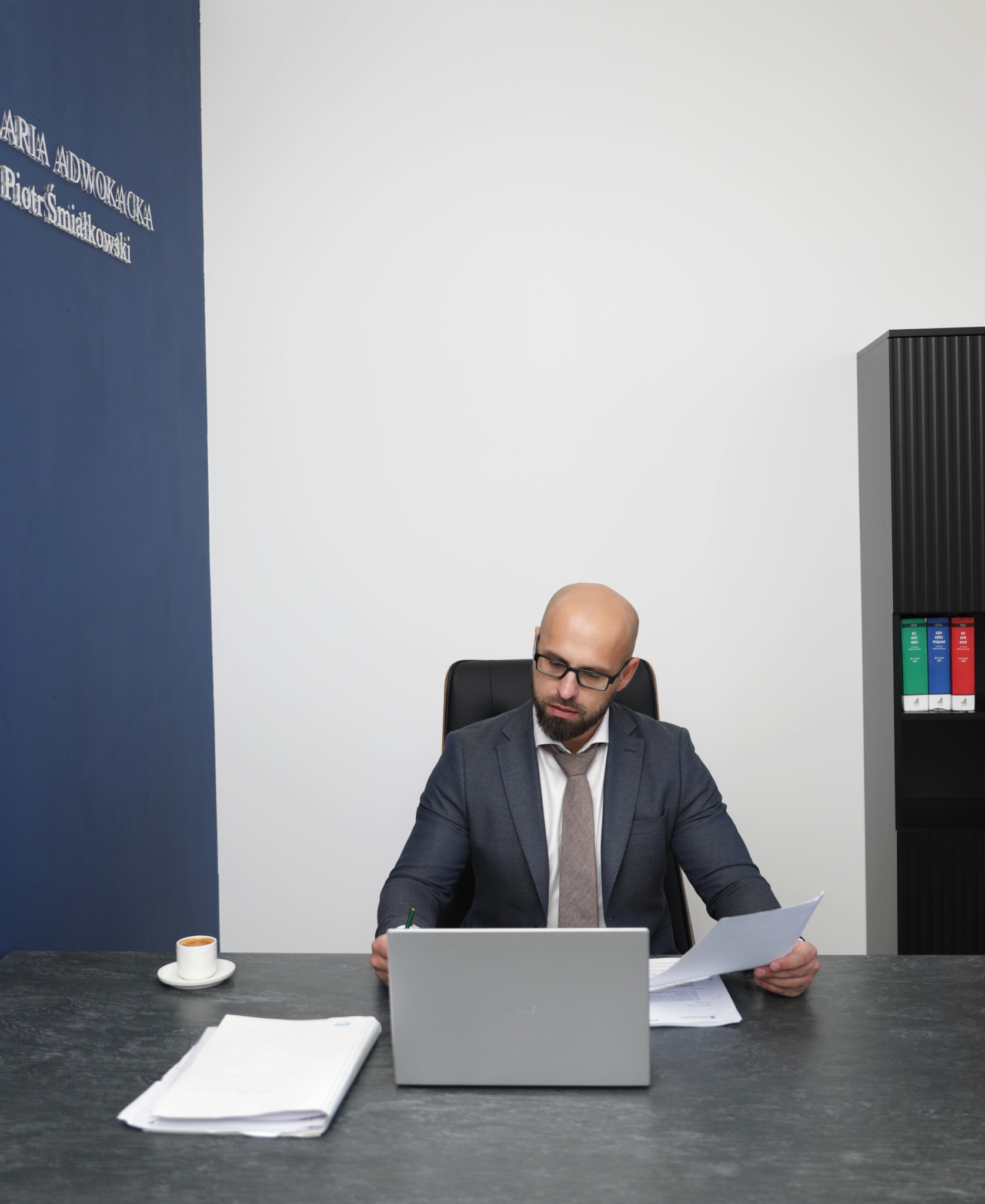 Adwokat w garniturze, z laptopem i dokumentami przy biurku w nowoczesnym biurze. Na ścianie widoczna nazwa kancelarii.