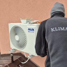 Klimator - Montaż klimatyzatora AUX na elewacji domu. Widoczny pracownik w polarze z logo firmy. Na klimatyzatorze leżą narzędzia.